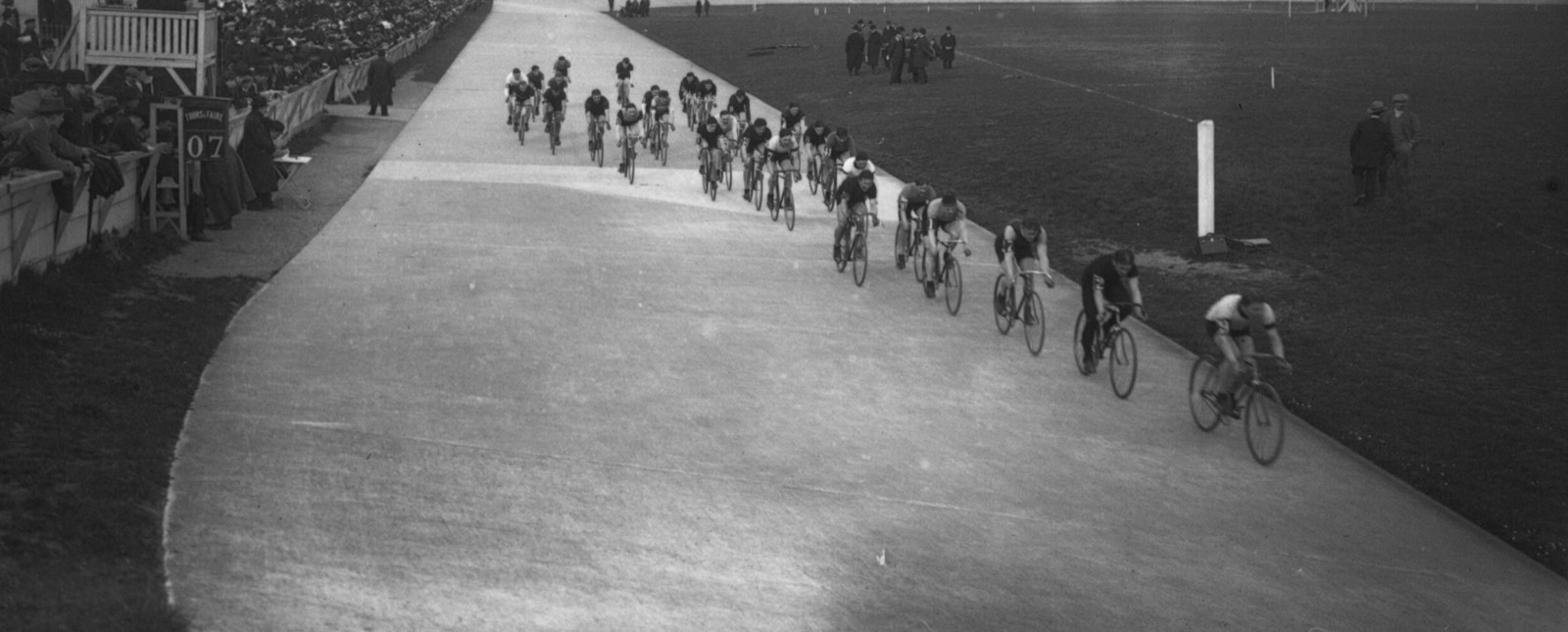 Fotografía de 1913, en blanco y negro, con un pelotón de ciclistas en la pista de cemento del Parc des Princes y público apoyado en la barrera