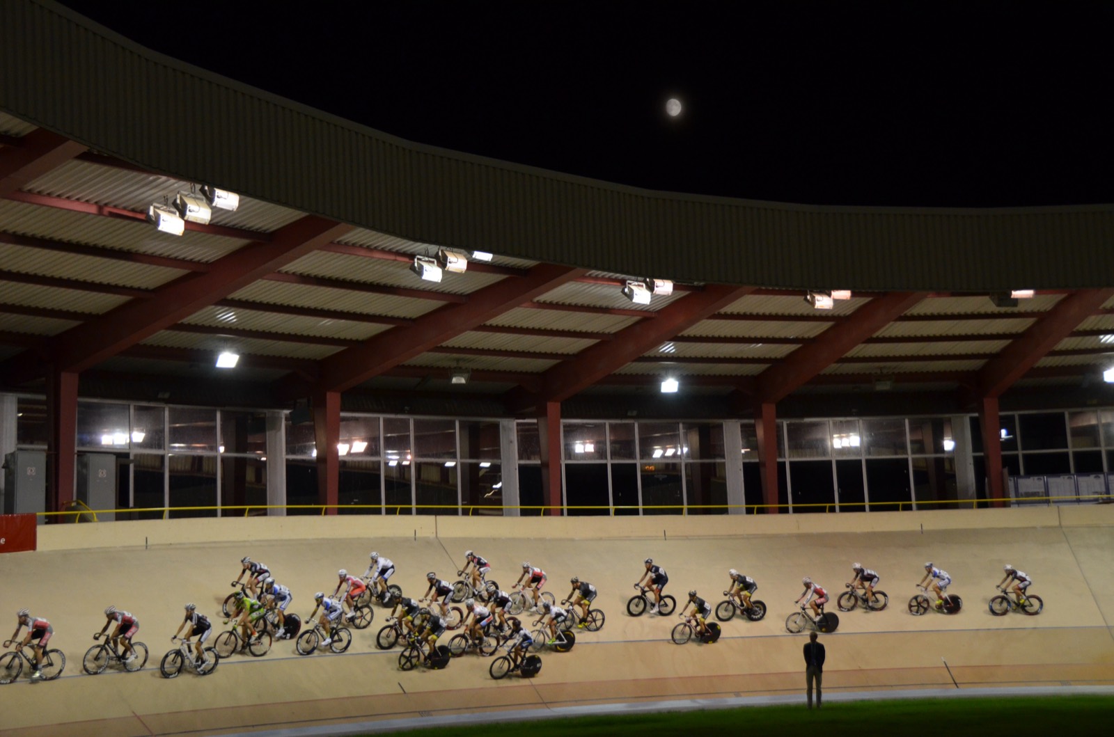 Pelotón de una carrera por puntos rodando de noche por la curva de un velódromo descubierto, bajo la cubierta iluminada de la grada y con la luna en el cielo