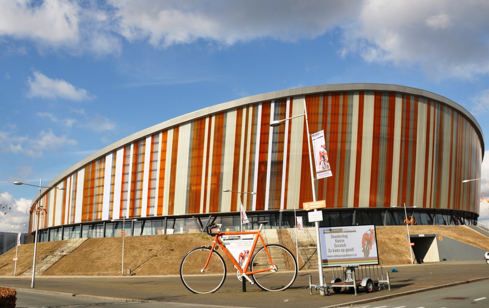 Fachada de franjas naranjas y blancas del Omnisport de Apeldoorn, con una bicicleta gigante de color naranja delante