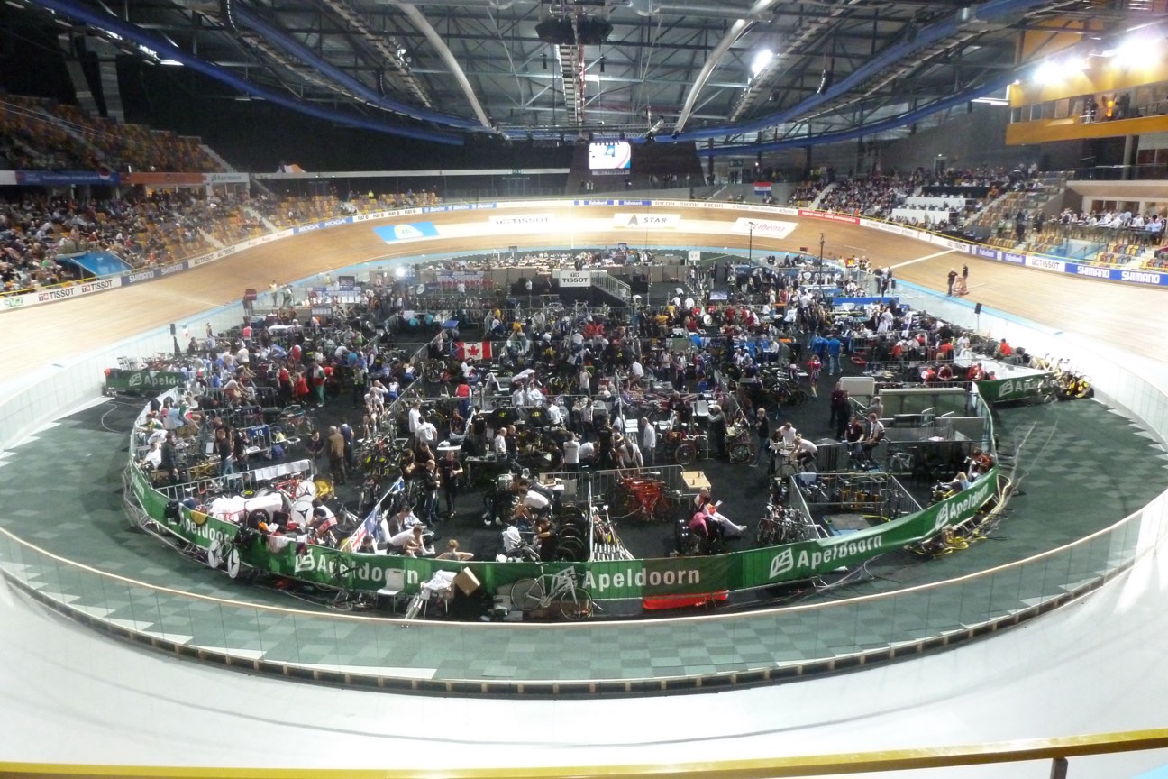 Interior del Omnisport de Apeldoorn desde la grada, con la pista de madera peraltada y, en el centro, los boxes de los equipos llenos de gente y bicicletas