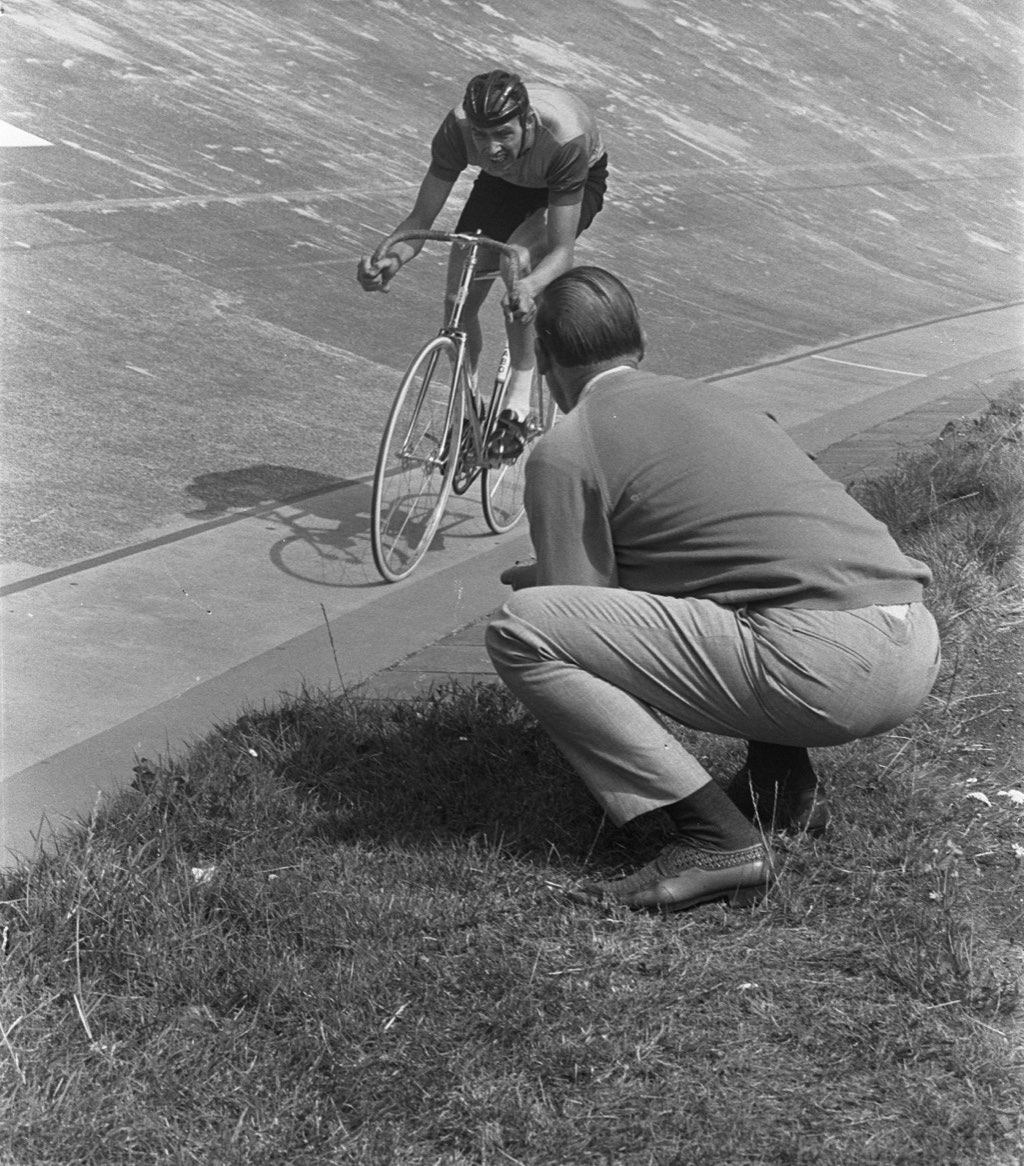 Un corredor sale de la curva mientras su entrenador, agachado en la hierba del interior, le da indicaciones, en una imagen en blanco y negro de 1967