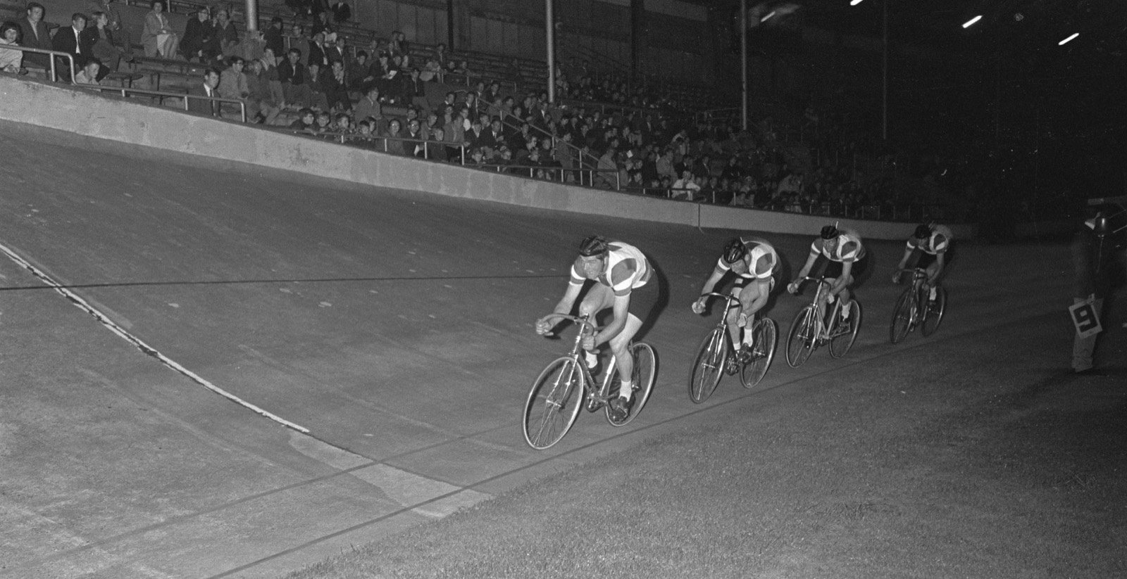 Un cuarteto de persecución, uno detrás de otro, entrando en la curva con la grada llena al fondo, en blanco y negro