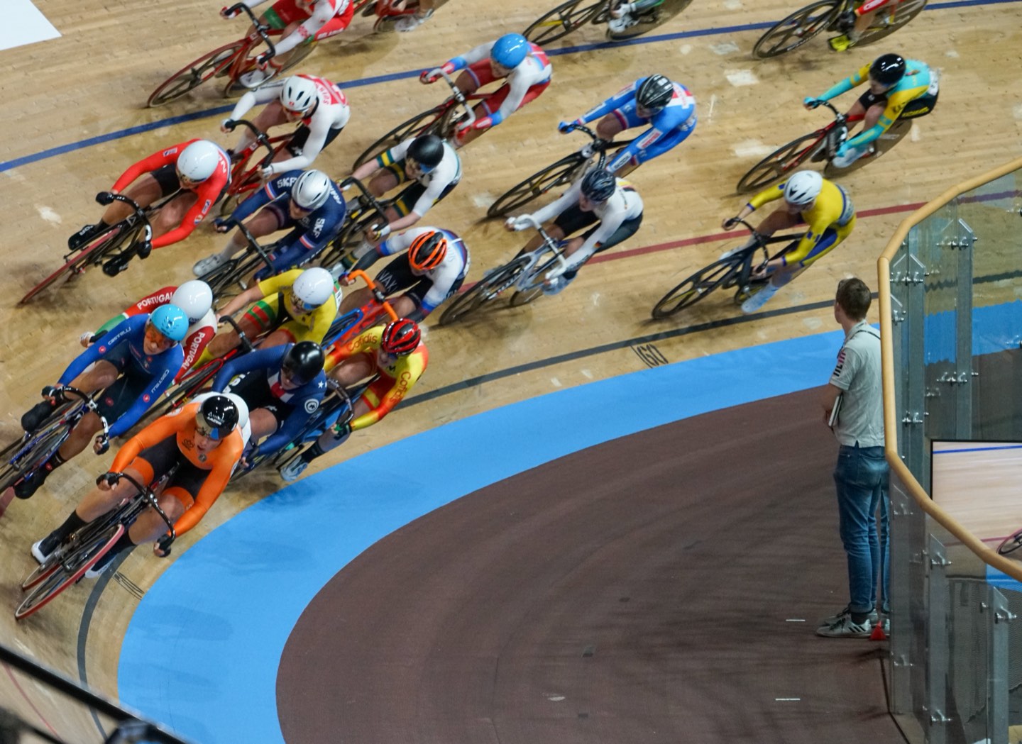 Pelotón de corredoras entrando en una curva, fotografiado desde lo alto de la grada, con la franja azul y una persona de pie dentro del óvalo