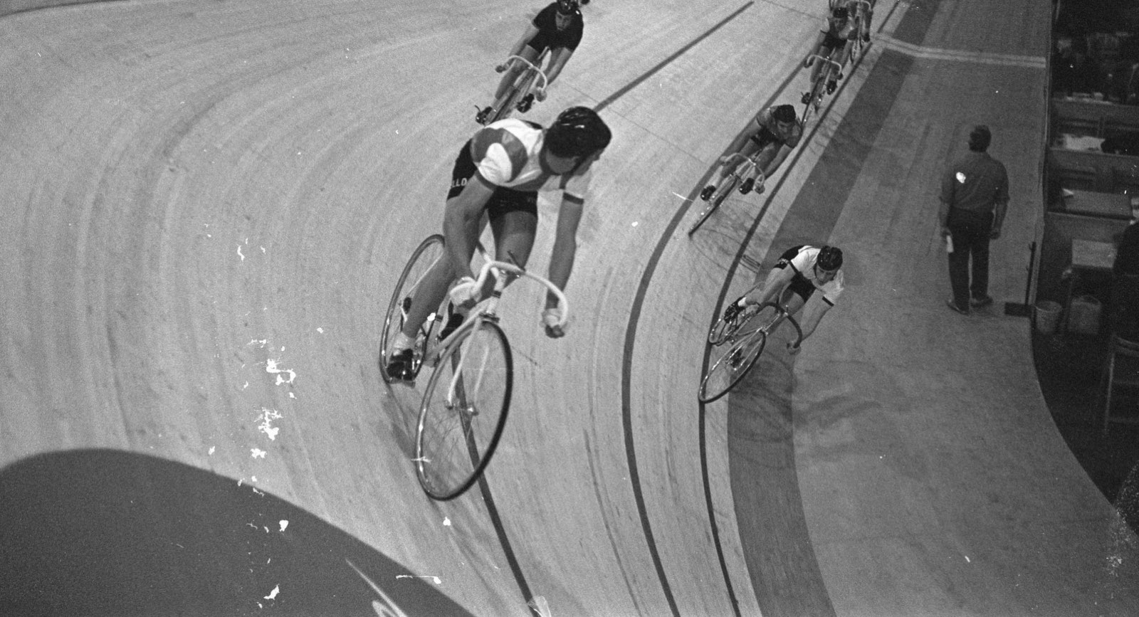 Corredores de una carrera de seis días bajando en fila por la curva peraltada de una pista de madera, vistos desde lo alto; foto en blanco y negro