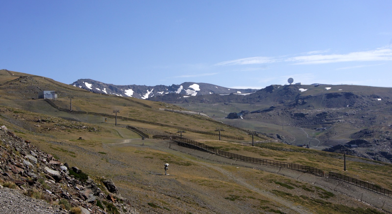 Laderas de Sierra Nevada en verano, casi sin nieve, con vallas y postes de remonte de la estación de esquí y las cumbres al fondo bajo un cielo despejado